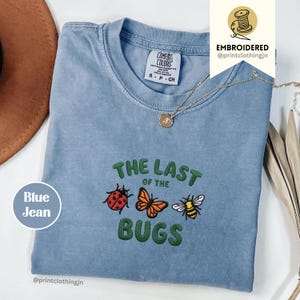 May include: A blue jean coloured t-shirt featuring an embroidered design of a ladybird, a butterfly, and a bee. The text "THE LAST OF THE BUGS" is embroidered in green above and below the insects. The shirt has a Comfort Colors brand neck label.