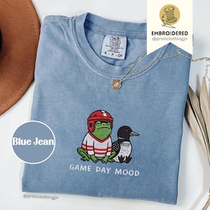 Könnte beinhalten: Blaues Jeans-T-Shirt mit einem gestickten Design, das einen Frosch mit Footballhelm und eine Eistaucher zeigt. Der Text "GAME DAY MOOD" steht darunter. Eine goldene Halskette liegt auf dem Shirt.
