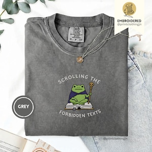 Könnte beinhalten: Graues T-Shirt mit dem Aufdruck „Scrolling the Forbidden Texts“ und einer Cartoon-Froschfigur mit Umhang und Stab. Das Shirt hat ein Comfort Colors-Etikett und eine Größenangabe.