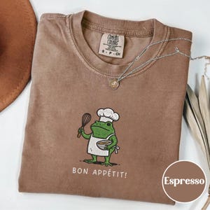Puede incluir: Camiseta color espresso con un dibujo de un chef rana sosteniendo un batidor y un cuenco, con gorro de chef y delantal. La frase "BON APPÉTIT!" está impresa debajo. Un collar plateado con un colgante está sobre la camiseta.
