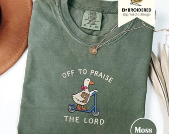 Puede incluir: Una camiseta verde musgo con la frase "OFF TO PRAISE THE LORD" bordada en blanco. El diseño presenta un pato montado en un patinete azul y sosteniendo una biblia. La camiseta tiene cuello redondo y mangas cortas. La marca "Comfort Colors" es visible.