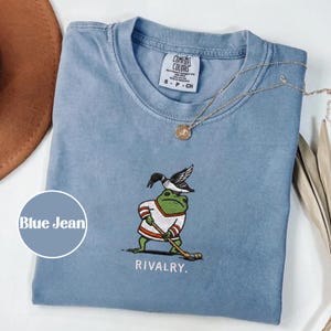 Könnte beinhalten: Hellblaues T-Shirt mit einer Stickerei einer Hockey spielenden Frosch, einer Eistaucher auf dem Kopf und dem Wort „RIVALRY“. Das T-Shirt hat einen Rundhalsausschnitt und kurze Ärmel. Eine goldene Halskette mit Anhänger liegt über dem T-Shirt.