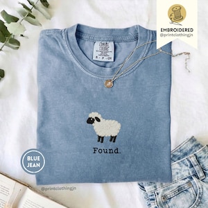 Puede incluir: Una camiseta azul de manga corta con cuello redondo que presenta un bordado de una oveja blanca con cara y patas negras en el centro del pecho. Debajo de la oveja, la palabra Found está bordada en negro. Un collar plateado descansa sobre el cuello.