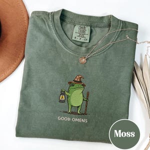 Puede incluir: Una camiseta verde musgo con una ilustración de una rana con sombrero de mago, sosteniendo una linterna y un bastón. El texto "GOOD OMENS" está debajo. Un collar dorado está sobre la camiseta.