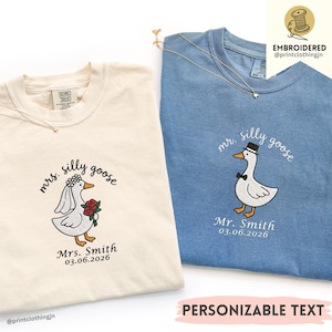 Könnte beinhalten: Ein cremefarbenes T-Shirt und ein blaues T-Shirt, beide mit gestickten Gänsemotiven. Das cremefarbene Shirt zeigt eine Gans mit Schleier und Rosen, beschriftet mit Mrs. Silly Goose, Mrs. Smith und 03.06.2026. Das blaue Shirt zeigt eine Gans mit Hut und Fliege, beschriftet mit Mr. Silly Goose, Mr. Smith und 03.06.2026.