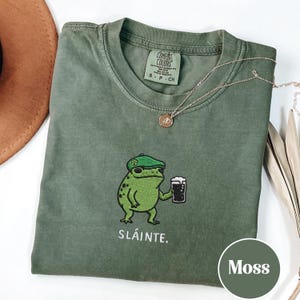 Peut inclure: T-shirt vert mousse avec un dessin de grenouille portant un béret vert et tenant une bière. Le mot "Sláinte" est imprimé en dessous. Un collier doré est posé sur le t-shirt.