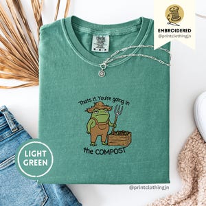 Op de afbeelding: Lichtgroen T-shirt met een geborduurd kikkerontwerp. De kikker draagt een overall en een strohoed, met een hooivork naast een compostbak. De tekst luidt: "That's it. You're going in the COMPOST".