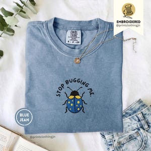 May include: A blue jean coloured t-shirt featuring an embroidered graphic of a blue beetle with yellow spots and the text STOP BUGGING ME. The shirt has a crew neckline and a Comfort Colors brand label on the inside collar. A thin silver chain necklace rests on the fabric.