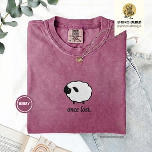 Könnte beinhalten: Ein beerenfarbenes T-Shirt mit Rundhalsausschnitt und einer Stickerei eines weißen Schafs mit schwarzem Gesicht und Ohren. Unter dem Schaf steht der Text "once lost.". Das T-Shirt hat ein Markenetikett am Kragen und ein "BERRY"-Label.