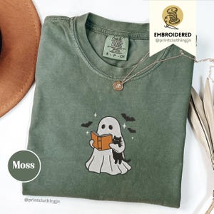Könnte beinhalten: Ein moosgrünes Kurzarm-T-Shirt mit einer Stickerei, die einen weißen Geist zeigt, der ein orangefarbenes Buch und eine schwarze Katze hält, umgeben von kleinen schwarzen Fledermäusen und Sternen. Das Shirt hat ein Comfort Colors Etikett.