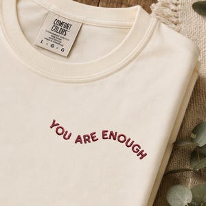 Könnte beinhalten: Ein cremefarbenes T-Shirt mit Rundhalsausschnitt und dem gestickten Text "YOU ARE ENOUGH" in dunkelrotem Garn. Das Shirt hat einen gerippten Halsausschnitt und ein weißes Innenetikett mit der Aufschrift "COMFORT COLORS" und der Größe "L G G".