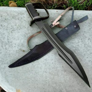May include: A black sword with a unique handle and a matching sheath. The blade has a dark finish with three parallel grooves. The handle features a finger guard. The sheath is made of dark leather.
