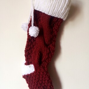 May include: A handmade Christmas stocking in a deep burgundy color with white accents. The stocking features a knitted design with a white cuff, toe, and heel. Two white pom-poms hang from the top. A festive holiday decoration.
