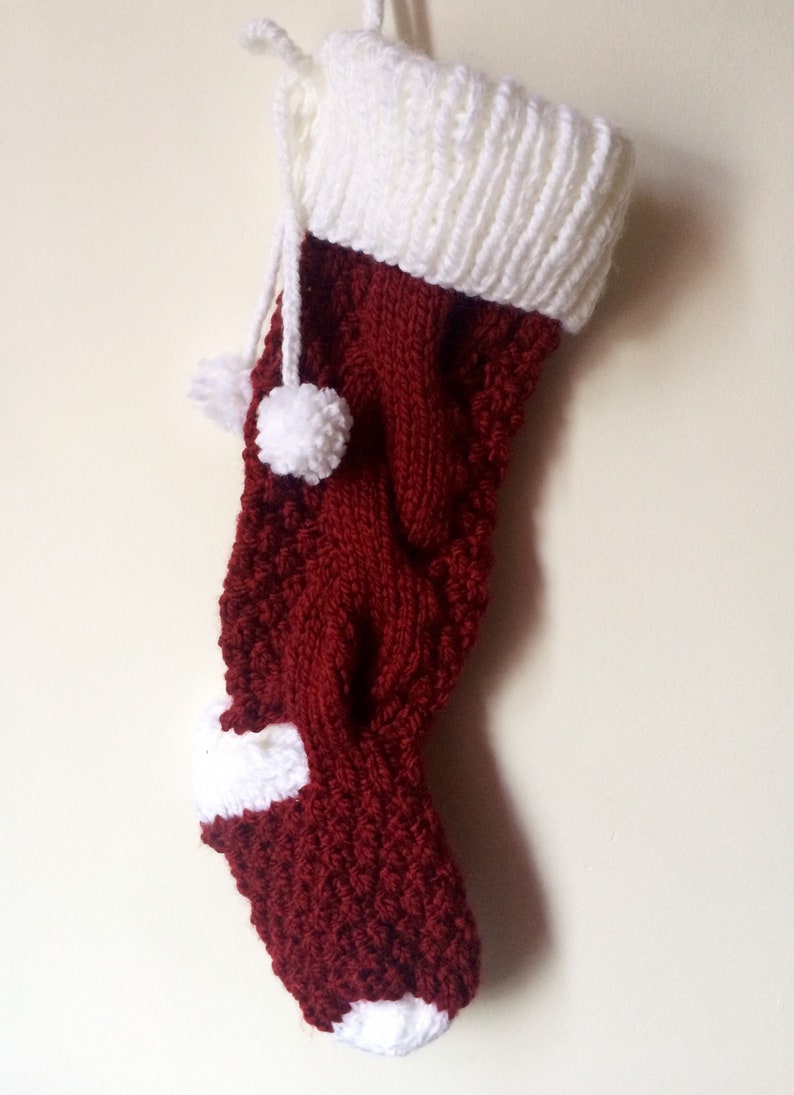 May include: A handmade Christmas stocking in a deep burgundy color with white accents. The stocking features a knitted design with a white cuff, toe, and heel. Two white pom-poms hang from the top. A festive holiday decoration.
