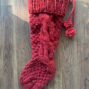 May include: A red knitted Christmas stocking with a cable knit design. The stocking has a ribbed cuff and two small pom-poms attached to a knitted cord. The stocking is made of a thick, textured yarn.