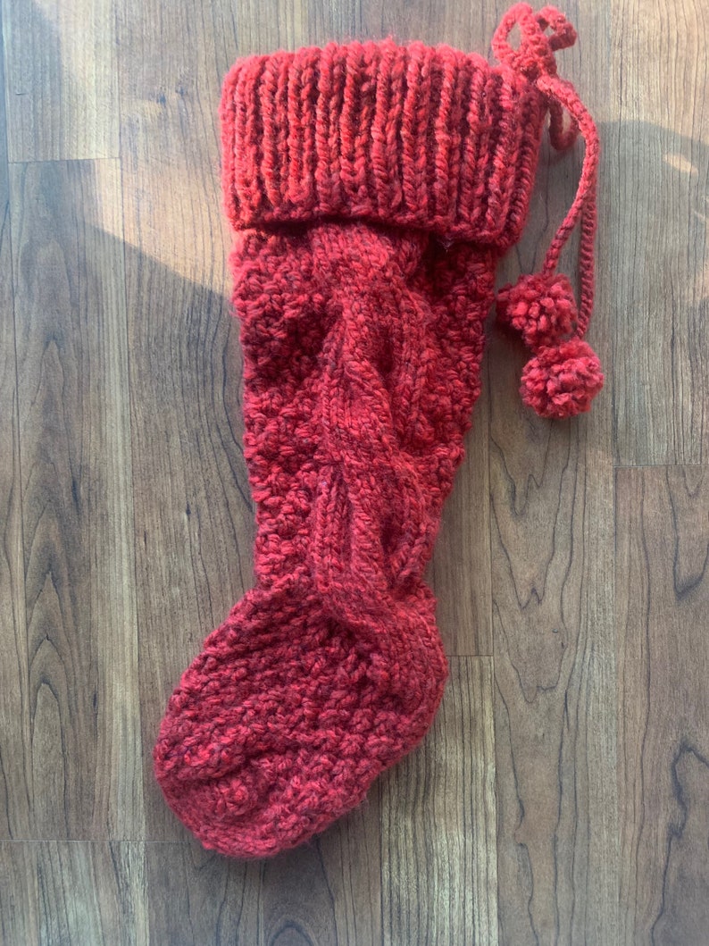 May include: A red knitted Christmas stocking with a cable knit design. The stocking has a ribbed cuff and two small pom-poms attached to a knitted cord. The stocking is made of a thick, textured yarn.