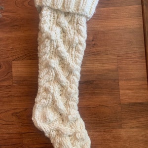 May include: Cream-colored, chunky knit Christmas stocking with a cable knit design. The top of the stocking is folded over, creating a ribbed cuff. The stocking is made of thick yarn and has a textured appearance.