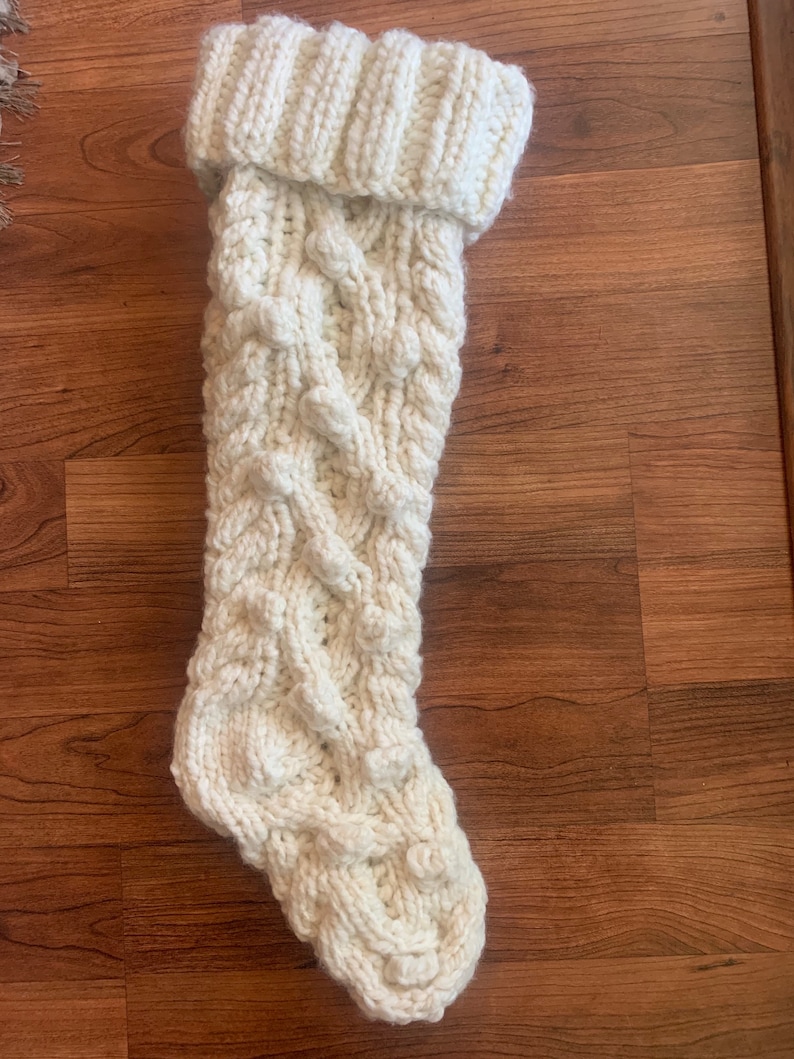 May include: Cream-colored, chunky knit Christmas stocking with a cable knit design. The top of the stocking is folded over, creating a ribbed cuff. The stocking is made of thick yarn and has a textured appearance.