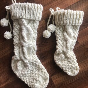 May include: Two off-white knitted Christmas stockings. The stockings have a cable knit design on the front and a ribbed cuff at the top. Each stocking has a loop for hanging and two white pom-poms attached.