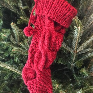 May include: A knitted Christmas stocking in a deep red hue, featuring a cable knit design and a ribbed cuff. The stocking is adorned with a small pom-pom and a loop for hanging, set against a backdrop of evergreen branches.