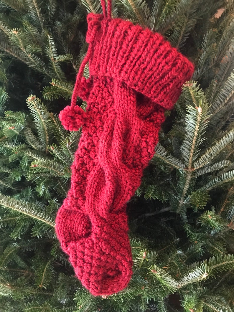 May include: A knitted Christmas stocking in a deep red hue, featuring a cable knit design and a ribbed cuff. The stocking is adorned with a small pom-pom and a loop for hanging, set against a backdrop of evergreen branches.