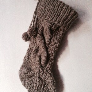 May include: A brown knitted Christmas stocking with a cable knit design and two small pom-poms. The stocking has a loop for hanging and is made of a soft, textured yarn. The overall style is rustic and cozy.