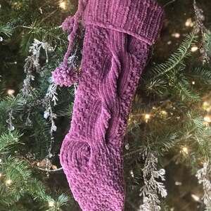 May include: A plush, mauve Christmas stocking with a cable knit design and pom-pom accents. The stocking is hanging on a Christmas tree with green needles and silver ornaments, creating a festive holiday scene.