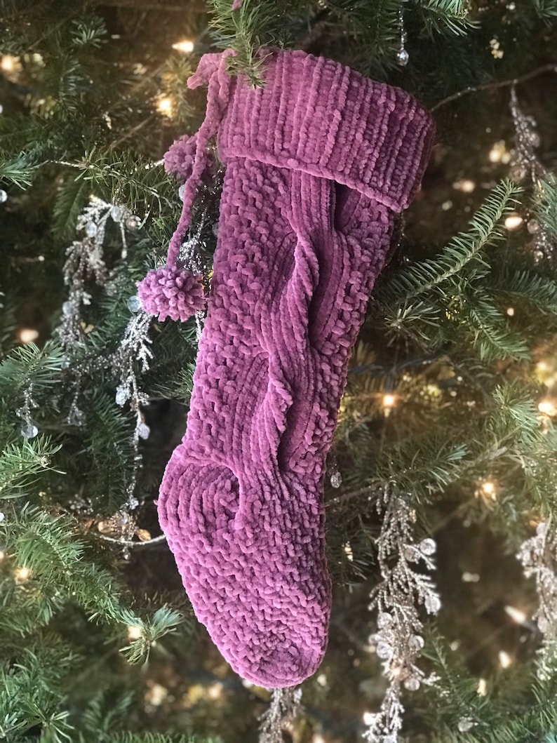 May include: A plush, mauve Christmas stocking with a cable knit design and pom-pom accents. The stocking is hanging on a Christmas tree with green needles and silver ornaments, creating a festive holiday scene.