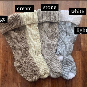 May include: A collection of five knitted Christmas stockings in various neutral colors: beige, cream, stone, white, and light gray. Each stocking features a cable knit design and a folded-over cuff. The stockings are displayed on a wooden surface.