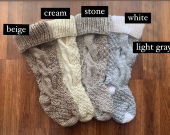 May include: A collection of five knitted Christmas stockings in various neutral colors: beige, cream, stone, white, and light gray. Each stocking features a cable knit design and a folded-over cuff. The stockings are displayed on a wooden surface.