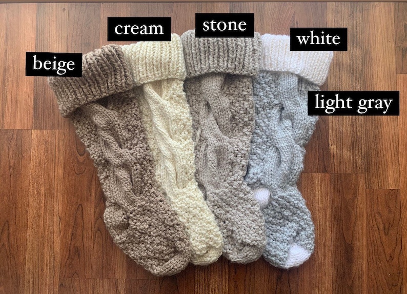 May include: A collection of five knitted Christmas stockings in various neutral colors: beige, cream, stone, white, and light gray. Each stocking features a cable knit design and a folded-over cuff. The stockings are displayed on a wooden surface.