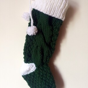 May include: A handmade Christmas stocking in dark green and white. The stocking is knitted with a cable knit pattern on the main body and a white cuff and toe. Two white pom-poms hang from the top. A loop is attached for hanging.