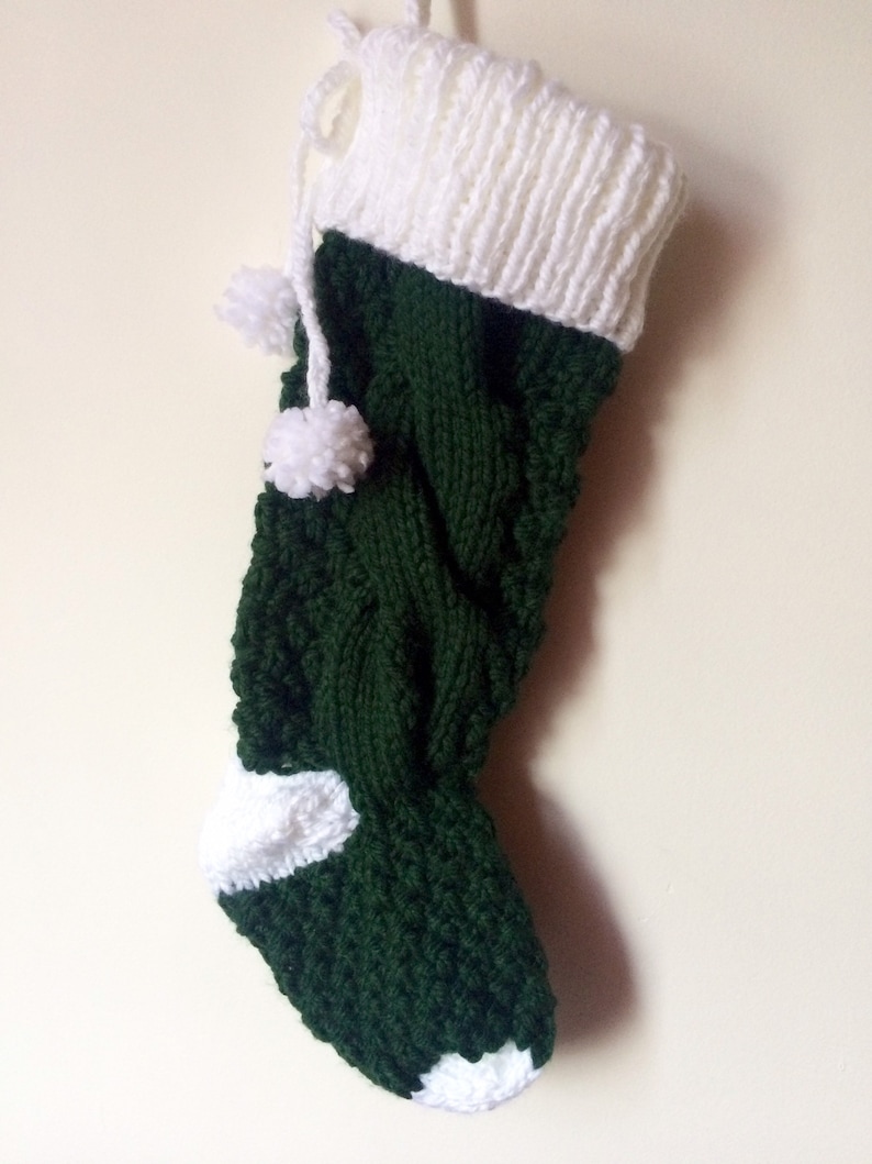 May include: A handmade Christmas stocking in dark green and white. The stocking is knitted with a cable knit pattern on the main body and a white cuff and toe. Two white pom-poms hang from the top. A loop is attached for hanging.