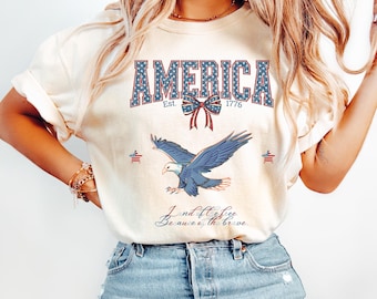 May include: A white t-shirt with a red, white, and blue graphic design. The graphic features the word "AMERICA" with stars, a red bow, and the text "Est. 1776". A blue eagle with red and white stars is flying above the text "Land of the free because of the brave."