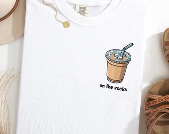 Peut inclure: T-shirt blanc avec un dessin animé d'un verre de café glacé avec une paille et l'inscription "on the rocks" en noir. Le t-shirt a un col rond et une étiquette Comfort Colors.