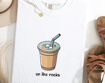 Peut inclure: T-shirt blanc avec une illustration de café glacé avec des glaçons et une paille. Les mots "on the rocks" sont imprimés en dessous en noir. Le t-shirt est posé à plat sur une surface blanche.