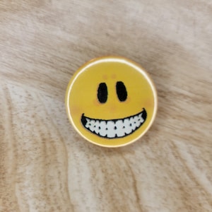 May include: A yellow smiley face button with black eyes and a wide, toothy grin. The button is round and has a glossy finish. The background is a light brown, wood-grain surface.