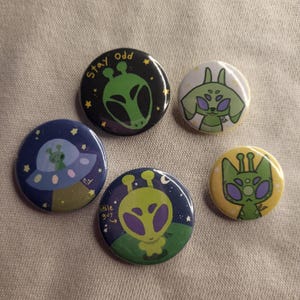 May include: Five round pinback buttons with alien illustrations. The buttons feature various designs, including an alien in a spaceship, an alien with the text "Stay Odd", and an alien with the text "Little Guy". The color palette includes green, blue, and yellow.