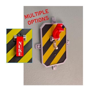 Könnte beinhalten: Eine Lichtschalterabdeckung mit einem gelb-schwarzen diagonalen Warnstreifenmuster. Die Abdeckung enthält eine rote, hochklappbare Sicherheitsabdeckung und einen zentralen Kippschalter. Ein Inset zeigt einen roten Hebel mit dem Text NUKE.