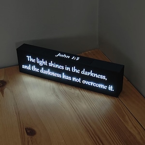 May include: A black, rectangular LED sign displaying the text "John 1:5 The light shines in the darkness, and the darkness has not overcome it." The sign is lit and placed on a wooden surface.