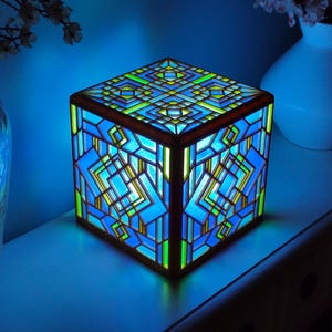 May include: A square lamp featuring a geometric stained glass design with vibrant blue, green, and yellow panels. The lamp emits a bright blue glow from within, highlighting the intricate lattice framework that forms a repeating pattern on each side.