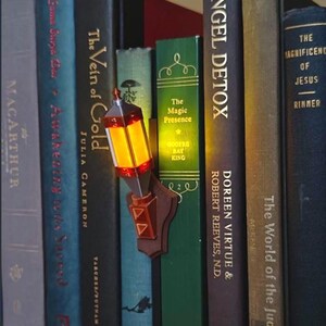 May include: A miniature wall-mounted lantern light fixture with a brown base and a glowing orange-yellow light, positioned between several hardcover books on a shelf. The lantern features a geometric design with red accents and a pointed top.