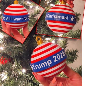 May include: Christmas ornament with red and white stripes and a blue band. The band has the text "All I want for Christmas!" and a star. The ornament has a gold-colored cap and is held in front of a Christmas tree.