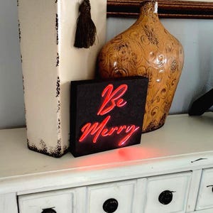 May include: A black square sign with the words "Be Merry" in red neon script. The sign is on a white table with a decorative vase and a framed picture in the background. The sign is lit up.