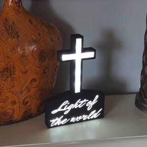 以下が含まれることがあります： 「Light of the world」の白い文字が入った、黒い十字架型の装飾ライト。十字架は明るい白い光を放っています。十字架の両側には2つの装飾的な花瓶があります。