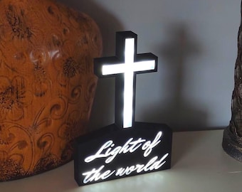 May include: A black cross-shaped decorative light with the words "Light of the world" in white script. The cross has a bright white glow. Two decorative vases are on either side of the cross.