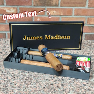 May include: A black cigar box with gold trim and the name "James Madison" in gold lettering. The box is open, revealing cigars, a lighter, and keys. The cigars are brown with colorful bands.