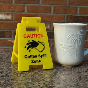 May include: A yellow "Coffee Spill Zone" caution sign with a black coffee spill graphic and the word "CAUTION" in red. A white mug with palm tree designs is next to the sign.