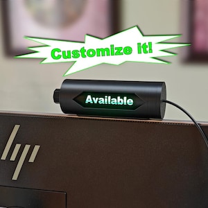 May include: A black cylindrical status indicator light mounted on top of a computer monitor. The device features a central hexagonal cutout displaying the illuminated green text "Available" and includes a black power cable extending from the side.