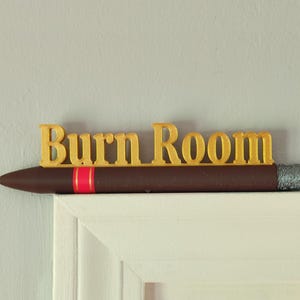 Può includere: Un cartello decorativo con la scritta "Burn Room" in lettere dorate. Il cartello ha la forma di un sigaro, con un corpo marrone, una fascia rossa e una punta argentata. Il cartello è posto su una superficie bianca.